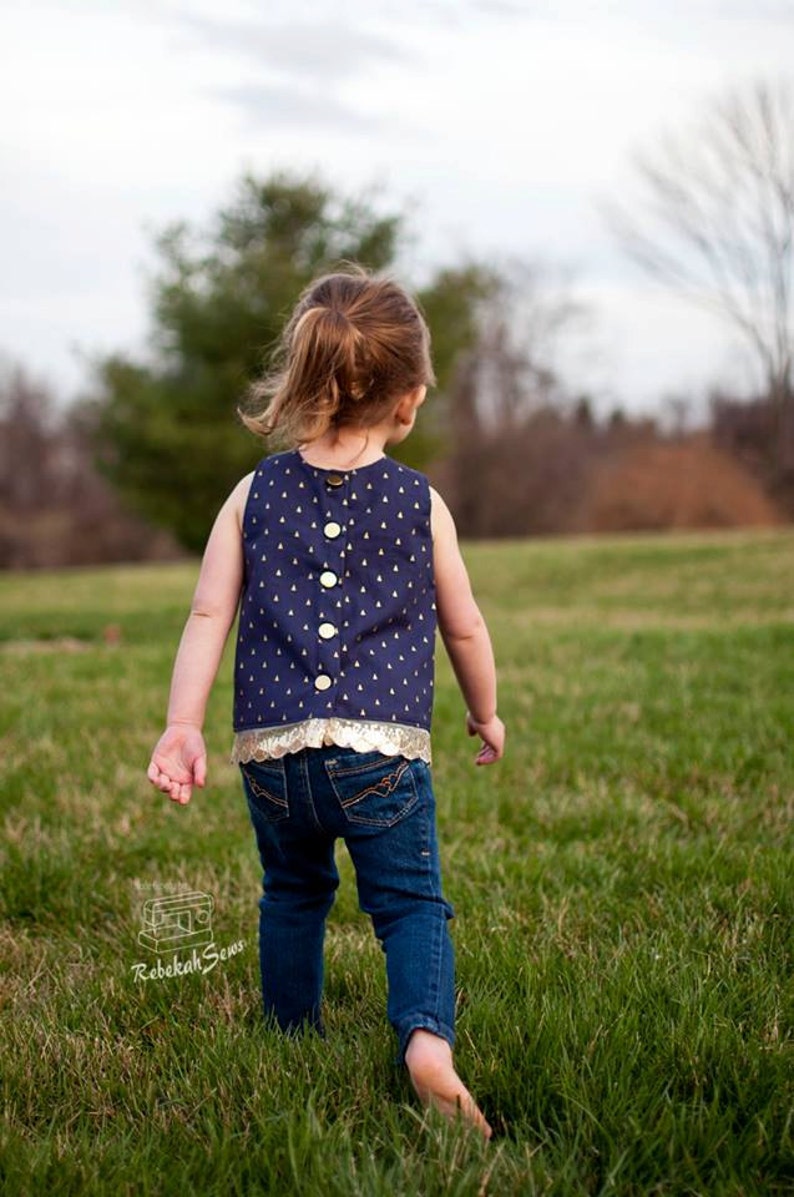 May include: A young person wearing a navy blue sleeveless top with gold accents and a gold button closure. The top has a white and gold pattern. The person is wearing blue jeans and is walking barefoot on green grass.