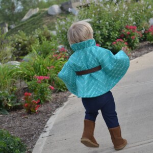 Puede incluir: Un ni&ntilde;o peque&ntilde;o con una capa de color turquesa y blanco con un cintur&oacute;n marr&oacute;n. El ni&ntilde;o lleva pantalones azules oscuros y botas marrones.