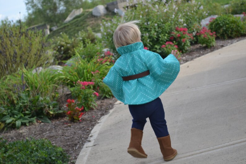 Puede incluir: Un ni&ntilde;o peque&ntilde;o con una capa de color turquesa y blanco con un cintur&oacute;n marr&oacute;n. El ni&ntilde;o lleva pantalones azules oscuros y botas marrones.
