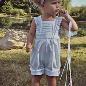Peut inclure: Une jeune enfant porte un bloomer bleu clair à pois blancs et bordé de dentelle, tenant des rubans et des ballons colorés. Le bloomer a une encolure carrée et des boutons. L'enfant porte un bandeau assorti et des chaussures bleu foncé.