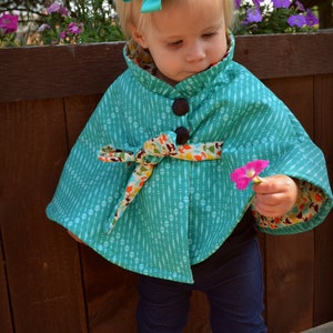 Puede incluir: Una ni&ntilde;a peque&ntilde;a con una capa azul turquesa con un ribete floral de colores y un lazo en el pelo. La capa tiene un cierre de bot&oacute;n y un lazo en la cintura. La ni&ntilde;a lleva pantalones vaqueros azules y botas marrones.
