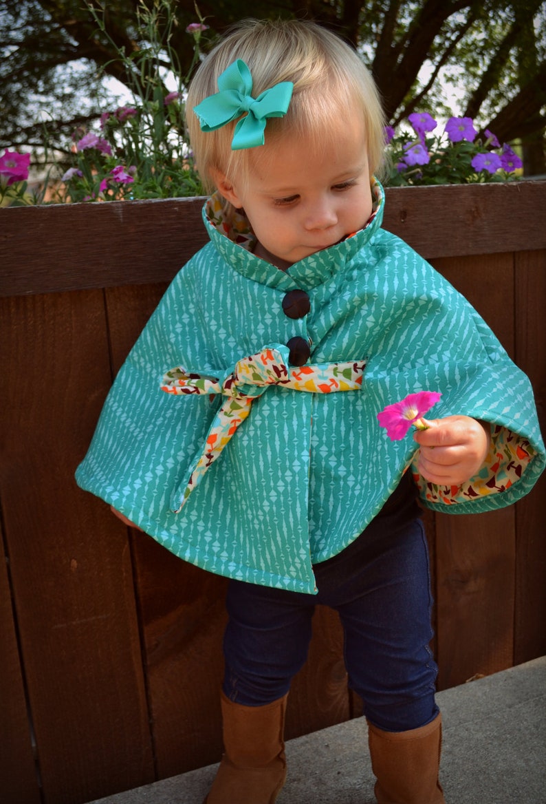 Puede incluir: Una ni&ntilde;a peque&ntilde;a con una capa azul turquesa con un ribete floral de colores y un lazo en el pelo. La capa tiene un cierre de bot&oacute;n y un lazo en la cintura. La ni&ntilde;a lleva pantalones vaqueros azules y botas marrones.