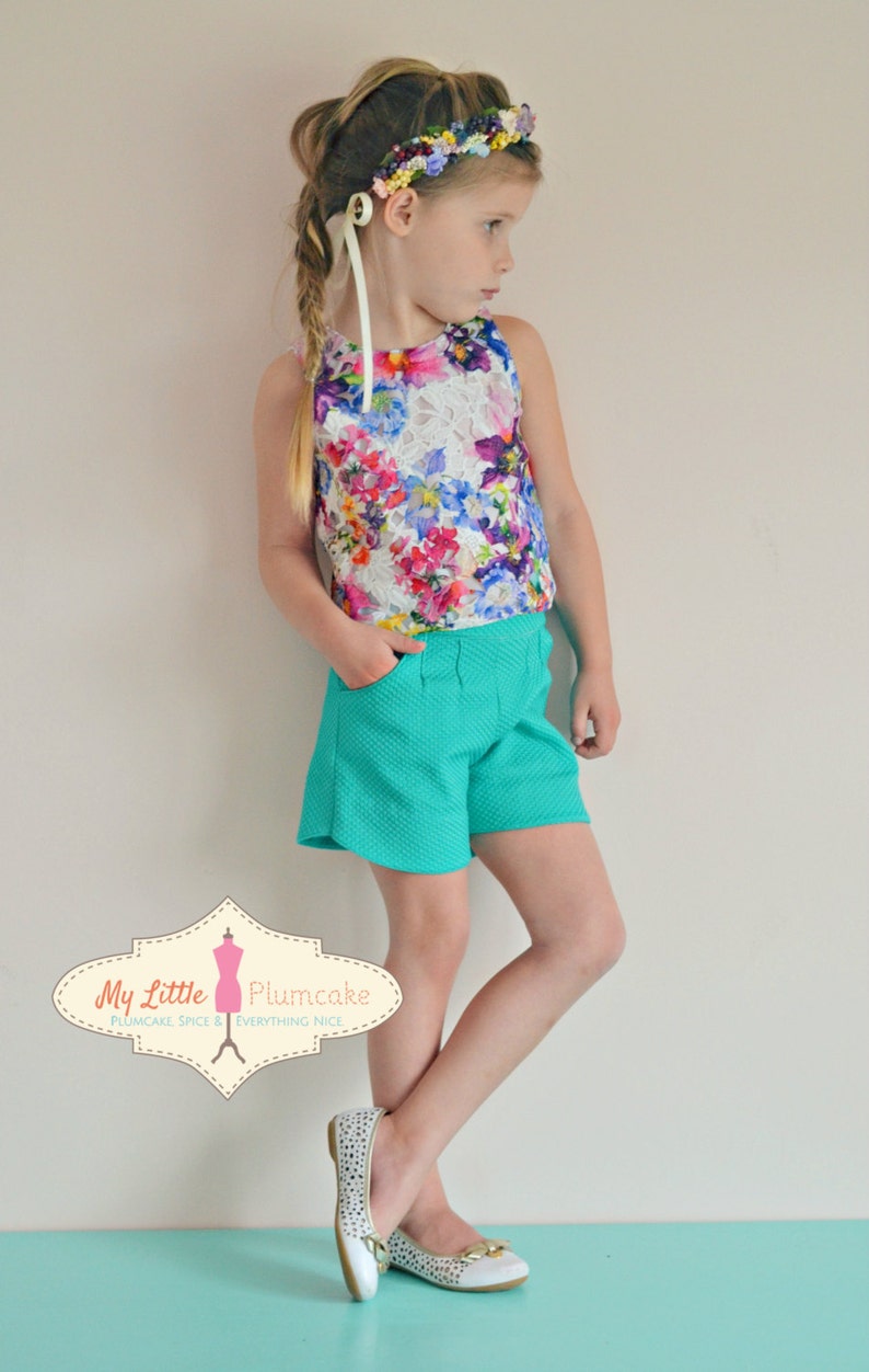 May include: A young girl wearing a floral print sleeveless top, teal shorts, and white shoes with gold accents. She is wearing a flower crown and has her hair in a braid. The image includes the text "My Little Plumcake Plumcake Spice & Everything Nice."