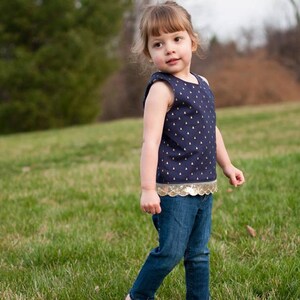 May include: A young girl wearing a navy blue sleeveless top with gold accents and a gold sequined trim. She is wearing blue jeans and is standing in a grassy field.