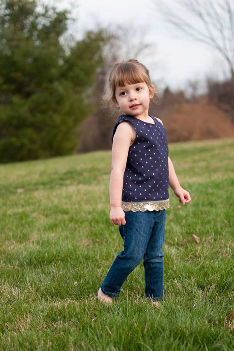 May include: A young girl wearing a navy blue sleeveless top with gold accents and a gold sequined trim. She is wearing blue jeans and is standing in a grassy field.