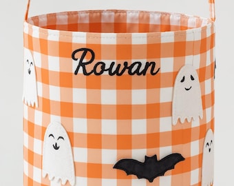 Puede incluir: Un cubo de tela cilíndrico con un patrón de cuadros vichy naranja y blanco y un asa fina naranja. El exterior está decorado con apliques de fantasmas blancos y un aplique de murciélago negro. El nombre Rowan está bordado en negro.