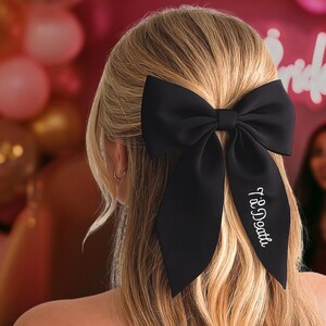 May include: A black satin hair bow with long tails, worn in the hair. The bow has the text "Til Death" embroidered in white script on one tail. The background includes pink balloons and the word "Bride" in neon.