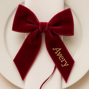 Puede incluir: Un lazo de terciopelo rojo oscuro con el nombre "Avery" bordado en hilo dorado. El lazo está colocado sobre una servilleta blanca, sobre un plato blanco. La textura y el color del lazo sugieren una ocasión festiva.