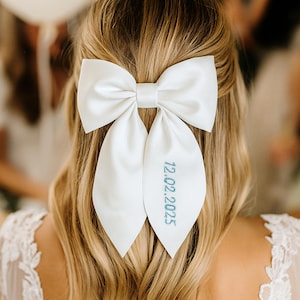 Può includere: Un fiocco per capelli in raso bianco con due lunghe code a punta. La coda destra è ricamata con la data 12.02.2025 in filo azzurro. Il fiocco è fissato sul retro di capelli biondi, mostrando una finitura in tessuto liscio e lucente.