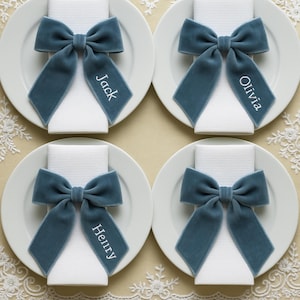 May include: Four white plates each hold a folded white napkin and a teal velvet bow. Each bow has a personalised name tag: Jack, Olivia, and Henry. The setting is on a cream-coloured surface with lace details.