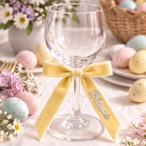 May include: A clear wine glass adorned with a yellow velvet ribbon tied in a bow. The ribbon has the name "Blaine" embroidered in a light blue script. The setting includes Easter eggs and floral arrangements.