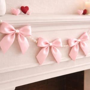 May include: A garland of three pink satin bows strung on a pearl strand. The bows are tied with a classic bow shape and are evenly spaced. The background includes small felt hearts and a white mantel.