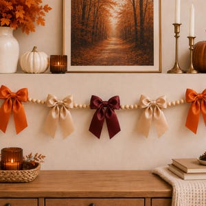 May include: A decorative garland featuring a string of light-colored wooden beads accented with five satin bows. The bows alternate in color, including two bright orange bows on the ends, two champagne-colored bows, and one deep burgundy bow in the center.