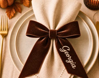 May include: A beige fabric napkin folded and secured with a dark brown velvet ribbon bow. The ribbon features the name Georgia embroidered in white script on one of the long, pointed tails. The napkin rests on a round, light-colored ceramic plate.