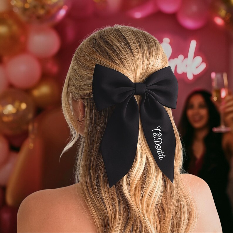 May include: A black satin hair bow with long tails, worn in the hair. The bow has the text "Til Death" embroidered in white script on one tail. The background includes pink balloons and the word "Bride" in neon.