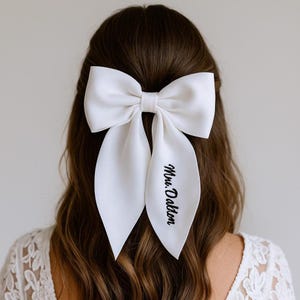 May include: A large white satin bow hair accessory. The bow is tied in the center and has long, flowing tails. The text "Mrs. Dalton" is printed in black script on one of the tails. The bow is worn in the hair.