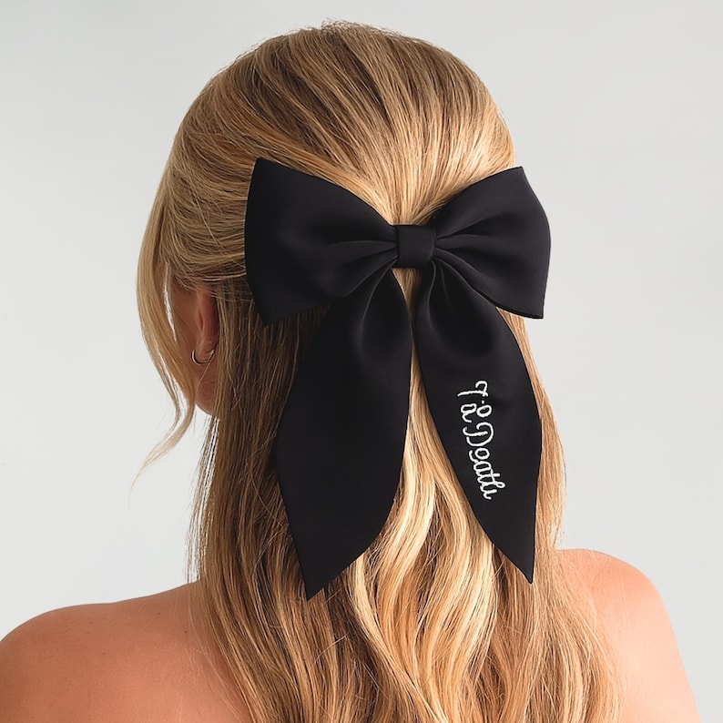 May include: A large black bow hair accessory with long, flowing tails. The bow is attached to the back of the head. The text "Til Death" is embroidered in white script on one of the tails. The hair is blonde.