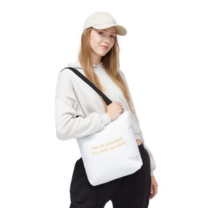 Può includere: Borsa tote bianca con tracolla nera. La borsa presenta il testo "Not an assistant. The whole operation." in lettere dorate. La borsa ha anche le lettere "OF" in oro.