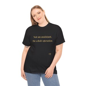 Può includere: T-shirt nero con testo dorato che recita "Not an assistant. The whole operation." La maglietta ha anche le iniziali "OF" in oro. La persona nell'immagine indossa jeans blu.