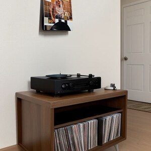 May include: A black turntable sits atop a brown wooden media console with silver legs. The console has open shelving filled with vinyl records. A framed album cover is mounted on the wall above the console.