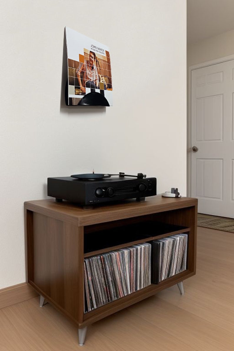 May include: A black turntable sits atop a brown wooden media console with silver legs. The console has open shelving filled with vinyl records. A framed album cover is mounted on the wall above the console.