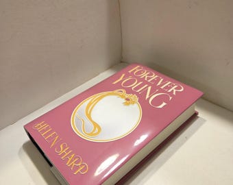 May include: A hardcover book titled "Forever Young" by Helen Sharp. The book has a pink cover with gold lettering and a circular design. The book is resting on a white surface.