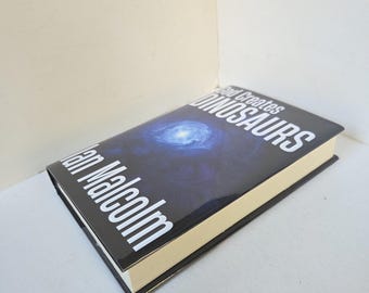May include: A hardcover book titled "God Creates Dinosaurs" by Ian Malcolm. The book has a dark blue cover with a galaxy image and white text. The book's pages are cream-colored, and the edges are black.