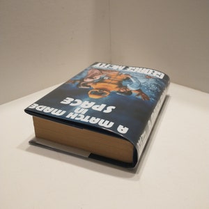 May include: A blue and white book titled "A Match in Space" by George Alec Effinger. The book cover features a photograph of two people in space suits.