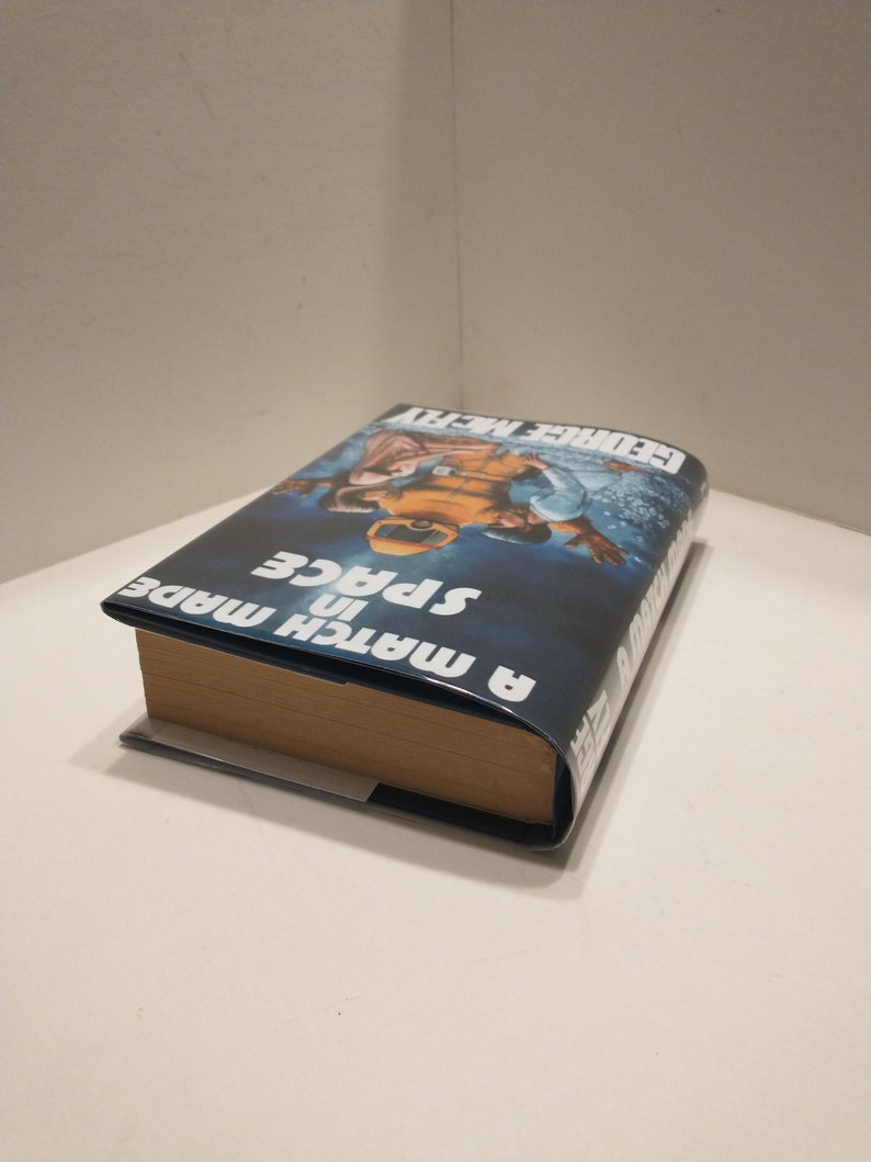 May include: A blue and white book titled "A Match in Space" by George Alec Effinger. The book cover features a photograph of two people in space suits.