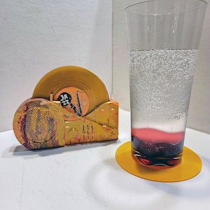 May include: A yellow record player-shaped coaster holder containing a yellow vinyl record with a label reading "JAZZ 22" and a saxophone graphic, alongside a tall clear glass of carbonated liquid with a dark red base resting on a yellow coaster.