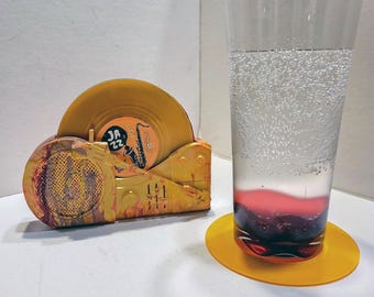 May include: A yellow record player-shaped coaster holder containing a yellow vinyl record with a label reading "JAZZ 22" and a saxophone graphic, alongside a tall clear glass of carbonated liquid with a dark red base resting on a yellow coaster.