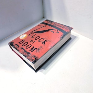 May include: A hardcover book titled "Clock of Doom" by Gale Weathers. The book has a red cover with black text and a distressed design. The book is resting on a white surface.