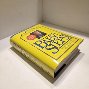 May include: A yellow book titled "BABY STEPS" with blue lettering. The book cover features a photo of a man and the author's name, Leo M. Marvin, M.D., Ph.D. The book is a guide to living life one step at a time.
