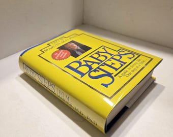 May include: A yellow book titled "BABY STEPS" with blue lettering. The book cover features a photo of a man and the author's name, Leo M. Marvin, M.D., Ph.D. The book is a guide to living life one step at a time.