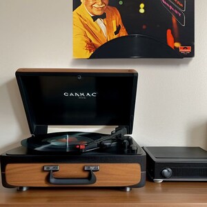 May include: A vintage-style record player with a wooden veneer and black accents, open to reveal a vinyl record. Above, a framed album cover with a portrait of a man in a yellow suit and the text "Fabulous Broadway".