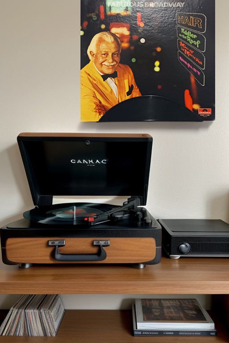 May include: A vintage-style record player with a wooden veneer and black accents, open to reveal a vinyl record. Above, a framed album cover with a portrait of a man in a yellow suit and the text "Fabulous Broadway".