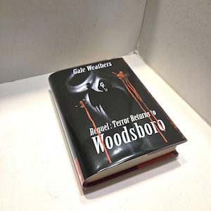 May include: A hardcover book titled "Requel: Terror Returns to Woodsboro" by Gale Weathers. The book has a black cover with a white graphic and red accents. The book is resting on a white surface.