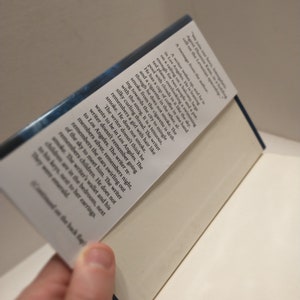 May include: A close-up of the back cover of a book with a blue background and white text. The text reads "We join our hero, tunergale Agent Phoenis Kane, in a heading cell on the Levanian spaceship. A message from the author: A writer wakes up, and he is in Los Angeles. He is leaning on a railing on a balcony, large enough for two people and a pot for plants, overlooking a swimming pool with clouds in it like the sky. He has a cup of coffee in one hand and a cigarette in the other, although he does not smoke. The smoke from the cigarette is drifting towards the city to mingle with the smog. It is a tenuous, silky curling thread. The writer remembers a girl with hair like smoke. He does not smoke. The writer doesn't think he wants to be in Los Angeles. The writer doesn't remember going to Los Angeles. The writer remembers silver, remembers night, remembers the stars swirling out of the sky to meet him. The writer remembers children. He does not smoke. The writer's wallet and his children are in the bedroom, next to his keys, next to her earrings. They were emerald..."