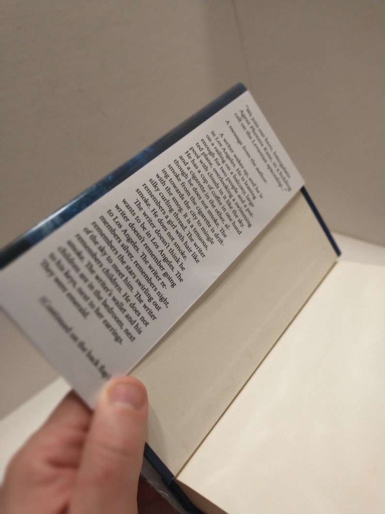 May include: A close-up of the back cover of a book with a blue background and white text. The text reads "We join our hero, tunergale Agent Phoenis Kane, in a heading cell on the Levanian spaceship. A message from the author: A writer wakes up, and he is in Los Angeles. He is leaning on a railing on a balcony, large enough for two people and a pot for plants, overlooking a swimming pool with clouds in it like the sky. He has a cup of coffee in one hand and a cigarette in the other, although he does not smoke. The smoke from the cigarette is drifting towards the city to mingle with the smog. It is a tenuous, silky curling thread. The writer remembers a girl with hair like smoke. He does not smoke. The writer doesn't think he wants to be in Los Angeles. The writer doesn't remember going to Los Angeles. The writer remembers silver, remembers night, remembers the stars swirling out of the sky to meet him. The writer remembers children. He does not smoke. The writer's wallet and his children are in the bedroom, next to his keys, next to her earrings. They were emerald..."