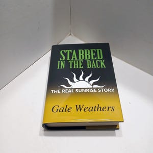 May include: A hardcover book titled "STABBED IN THE BACK" with a black cover and green text. The book features a sunrise graphic and the author's name, Gale Weathers, in gold. The book's spine is yellow.
