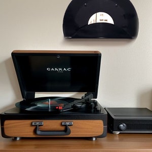 May include: A wooden and black record player with the brand name "CANNAC" on the front. A black record is playing on the turntable. A black record holder with a record is mounted on the wall above the record player.