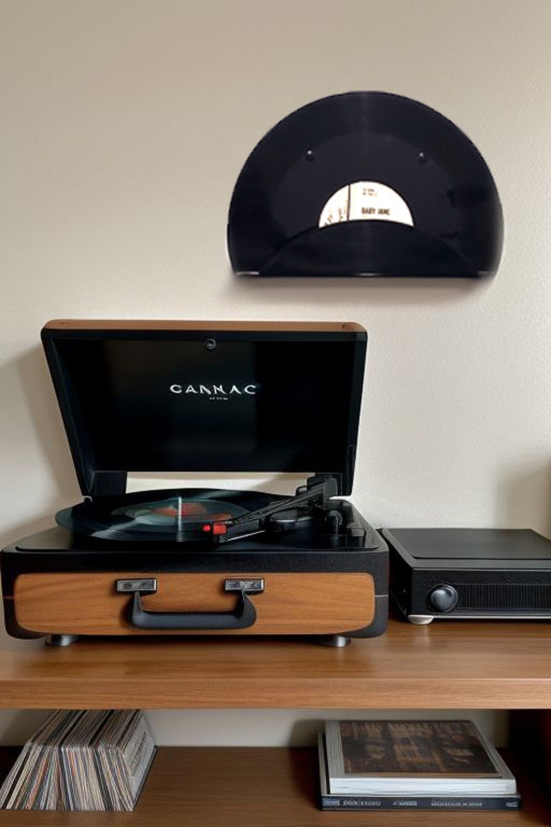 May include: A wooden and black record player with the brand name "CANNAC" on the front. A black record is playing on the turntable. A black record holder with a record is mounted on the wall above the record player.