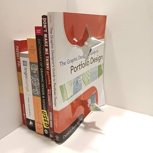 May include: A stack of books with a silver bookend shaped like a star. The top book is red and has the title "The Graphic Design Guide to Portfolio Design" on the cover. The bookend is holding the books in place.