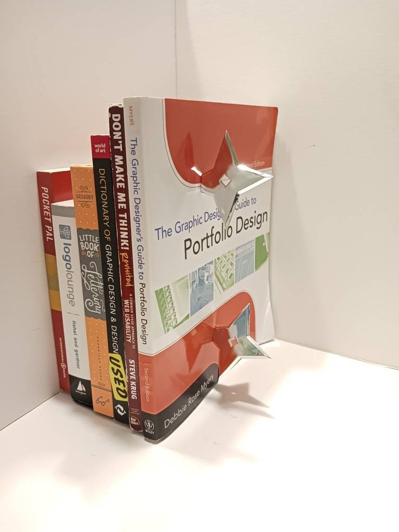 May include: A stack of books with a silver bookend shaped like a star. The top book is red and has the title "The Graphic Design Guide to Portfolio Design" on the cover. The bookend is holding the books in place.
