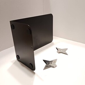 May include: A black metal bookend with four rubber feet on the bottom. Two silver throwing stars are on a white surface in front of the bookend.