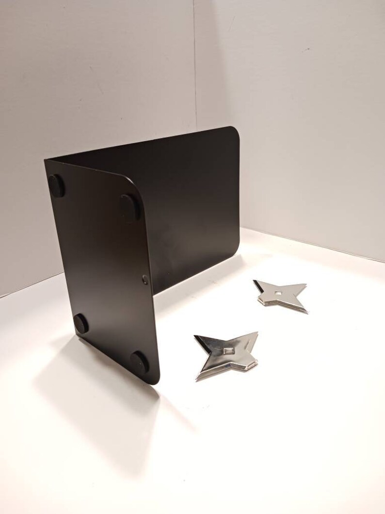 May include: A black metal bookend with four rubber feet on the bottom. Two silver throwing stars are on a white surface in front of the bookend.