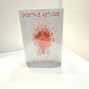 May include: A white rectangular product with the text "GHOSTFACE RETURNS" in red at the top and "GALE WEATHERS" at the bottom. A red and pink splatter design is in the center.
