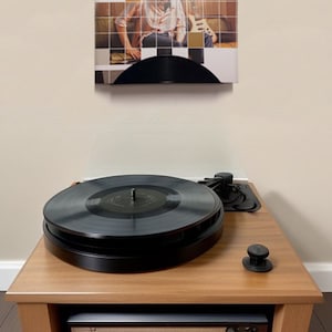 May include: A wooden record player with a black vinyl record on top. The record player is on a wooden stand with a shelf. Above the record player is a framed album cover that says "John Mayer Room for Squares".