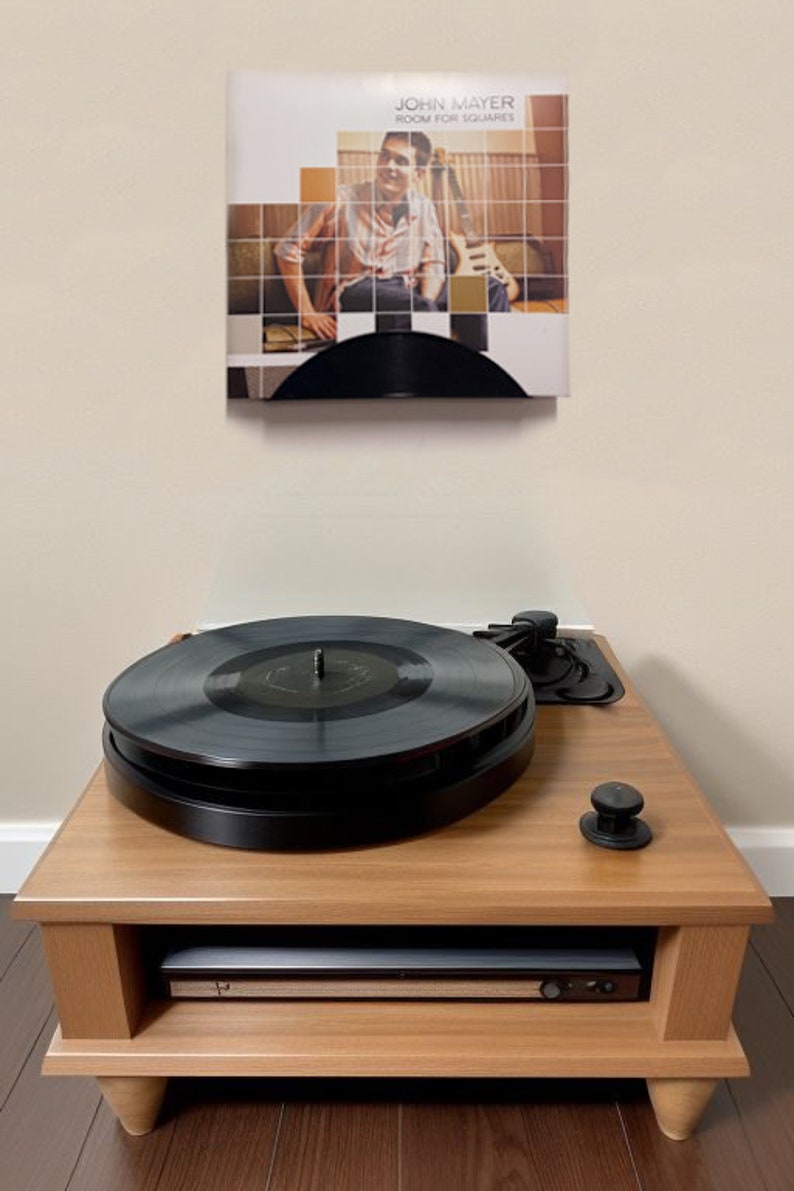 May include: A wooden record player with a black vinyl record on top. The record player is on a wooden stand with a shelf. Above the record player is a framed album cover that says "John Mayer Room for Squares".