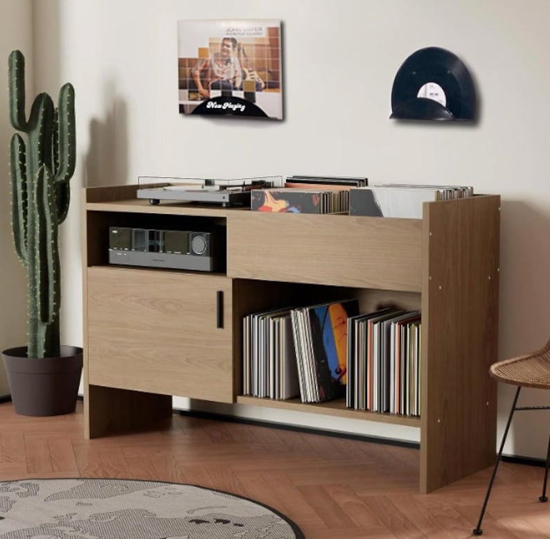 May include: A light oak record player console with a turntable, amplifier, and storage for vinyl records. A cactus and a John Mayer album are visible. The console has a modern design and is suitable for a living room or entertainment space.
